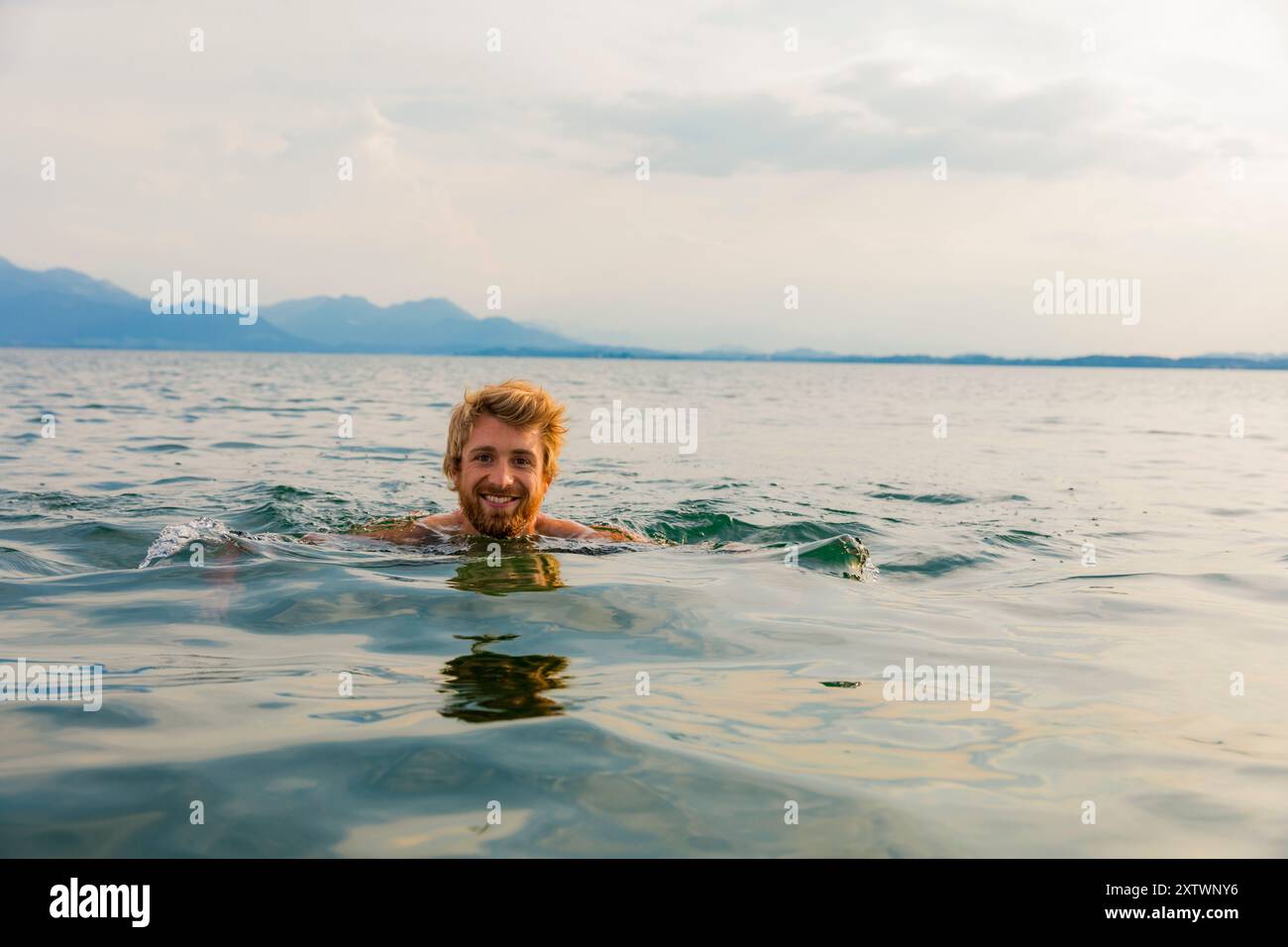 Uomo sorridente barbuto che nuota in un vasto e calmo lago con montagne in lontananza sotto un cielo soffice e nuvoloso. Foto Stock
