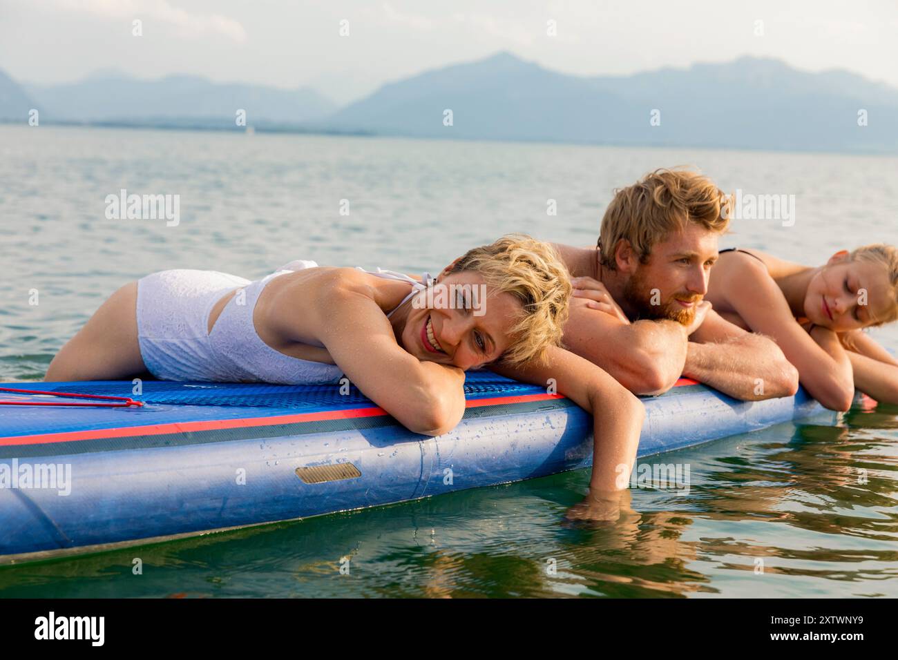 Tre amici si godono una giornata di sole mentre riposano su un paddleboard nelle calme acque del lago con montagne sullo sfondo. Foto Stock