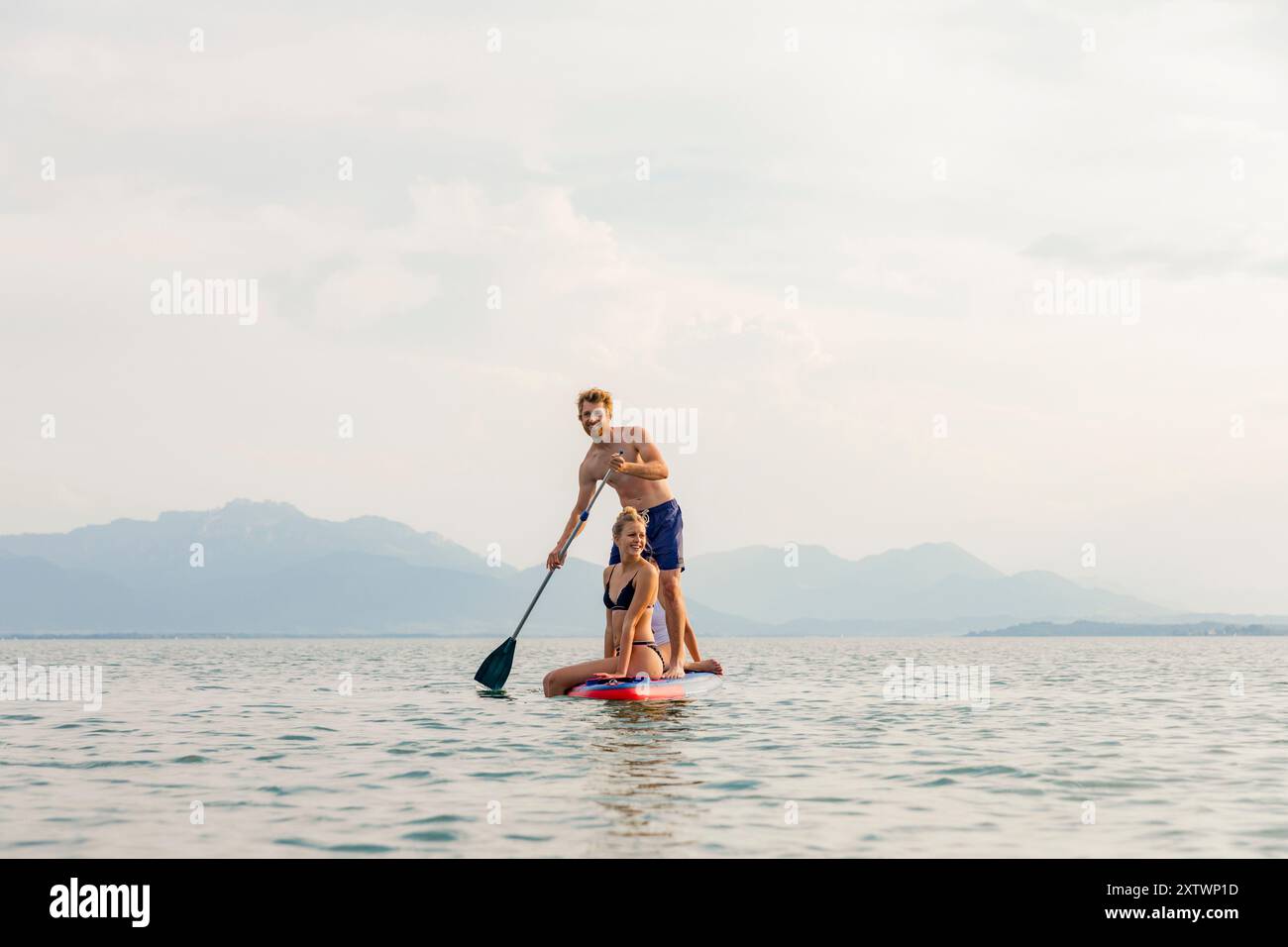 Un uomo e due bambini si divertono a fare paddleboard su un lago tranquillo con montagne sullo sfondo. Foto Stock