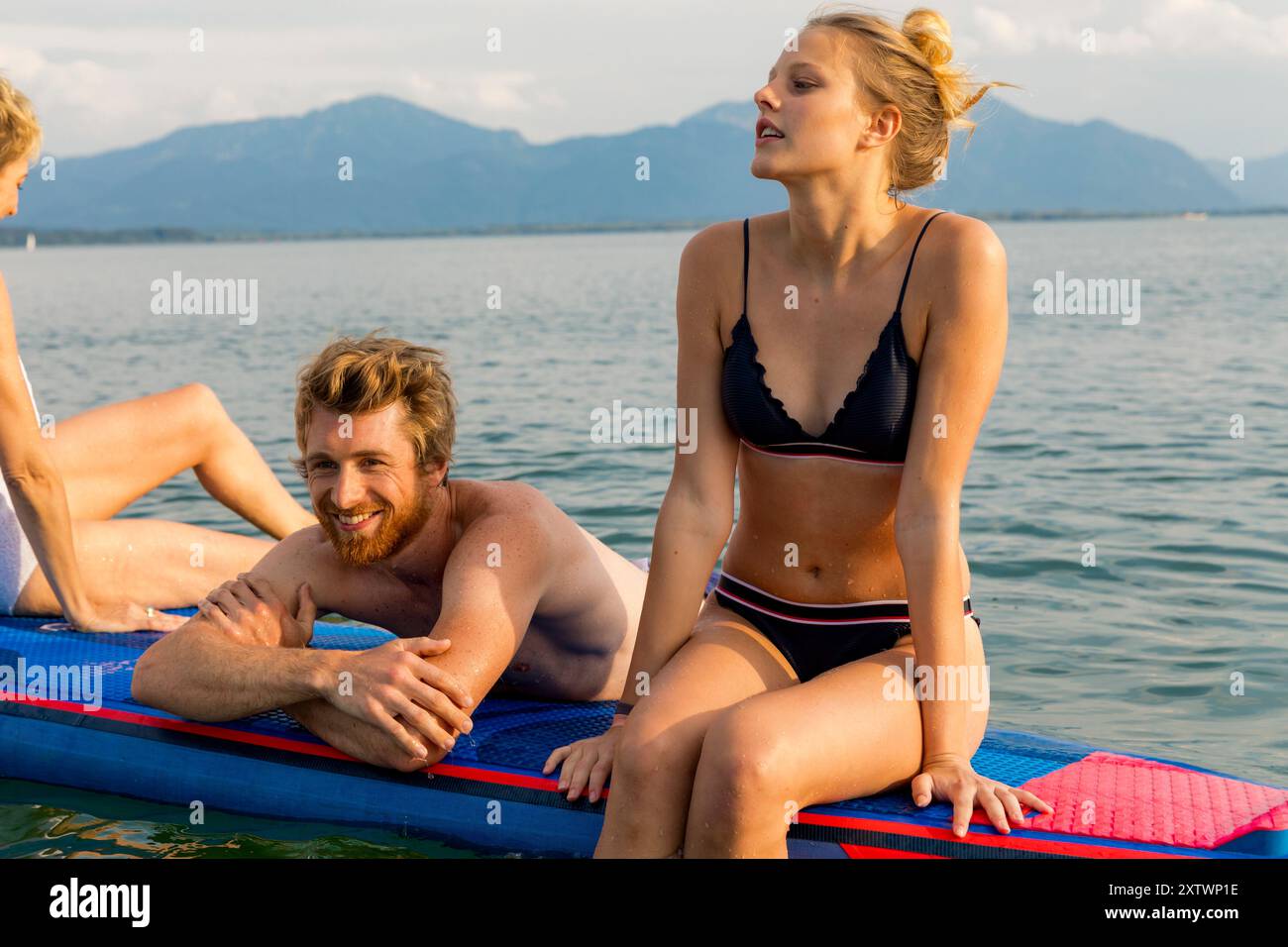 Due giovani adulti che si godono una giornata estiva su un tappeto galleggiante in un lago con montagne sullo sfondo. Foto Stock