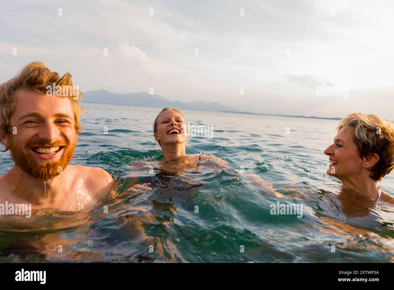 Tre persone gioiose che nuotano e ridono insieme nel mare con un cielo limpido sullo sfondo. Foto Stock