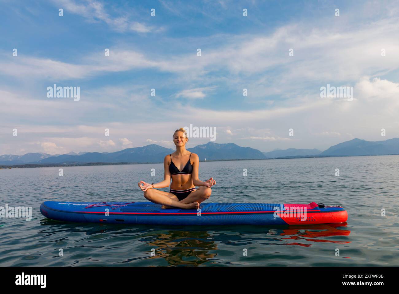 Una donna medita in posizione di loto su un paddleboard galleggiante su un lago calmo con montagne in lontananza. Foto Stock