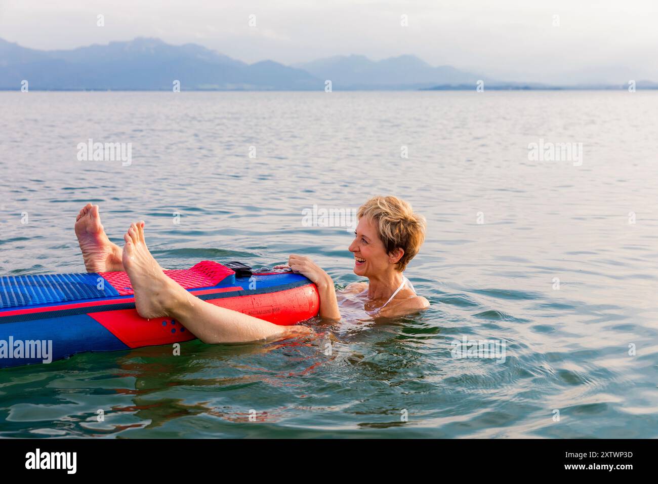 Una donna allegra con capelli corti che galleggia su un tappeto blu in un lago calmo con montagne in lontananza. Foto Stock