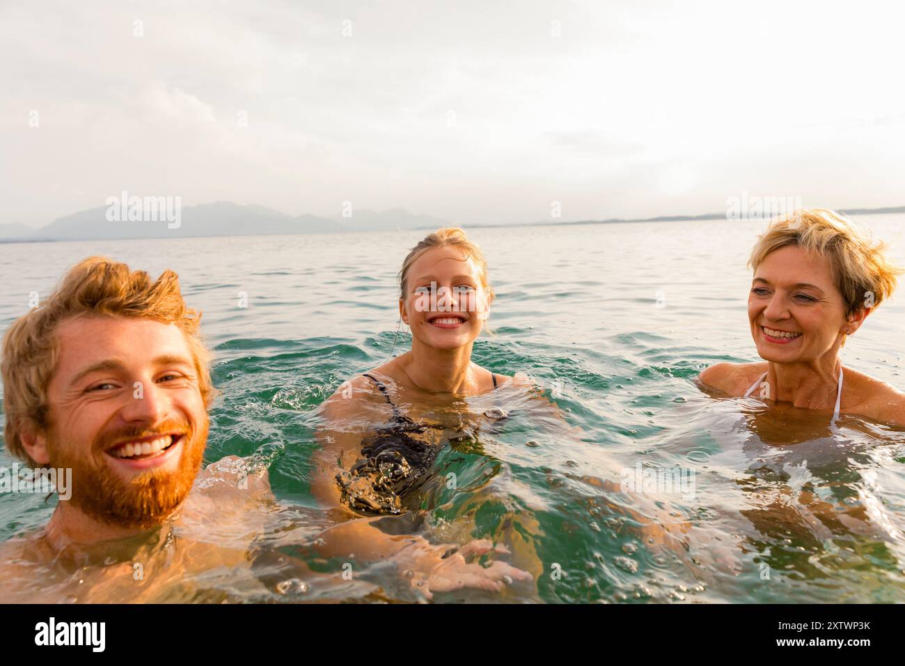 Tre persone nuotano gioiosamente in un ambiente aperto con montagne sullo sfondo al tramonto. Foto Stock