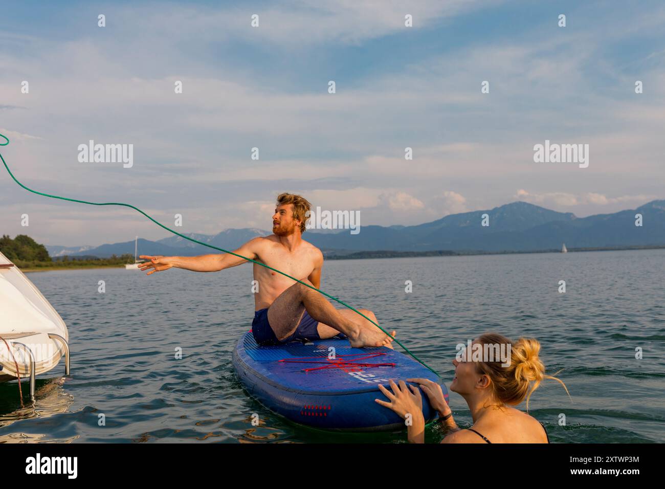 Uomo seduto su un wakeboard che riceve una corda di traino da una donna da una barca su un lago calmo con montagne sullo sfondo. Foto Stock