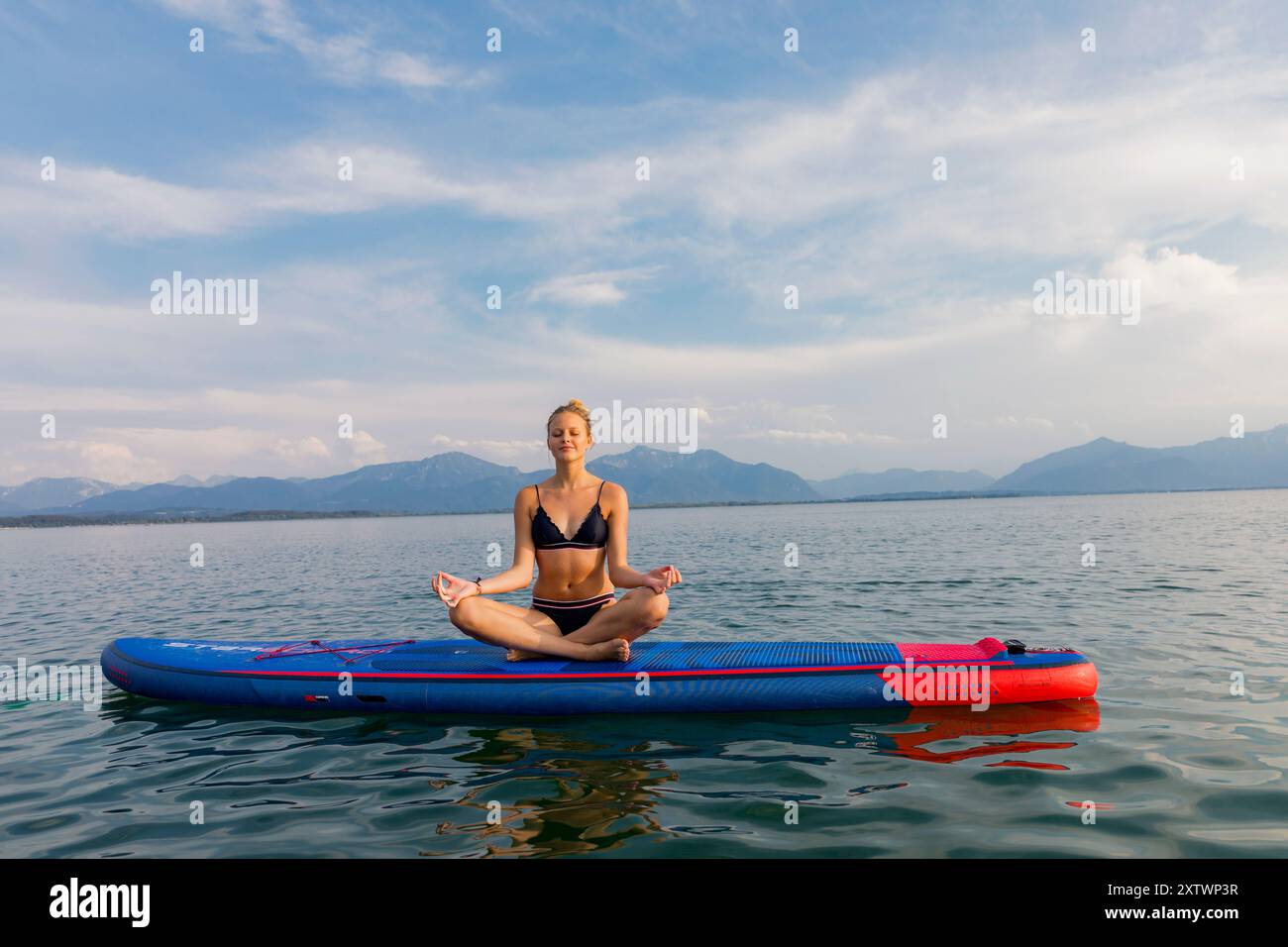 La donna medita in una tranquilla posa yoga su una tavola da paddleboard nelle acque calme di un lago con montagne sullo sfondo. Foto Stock