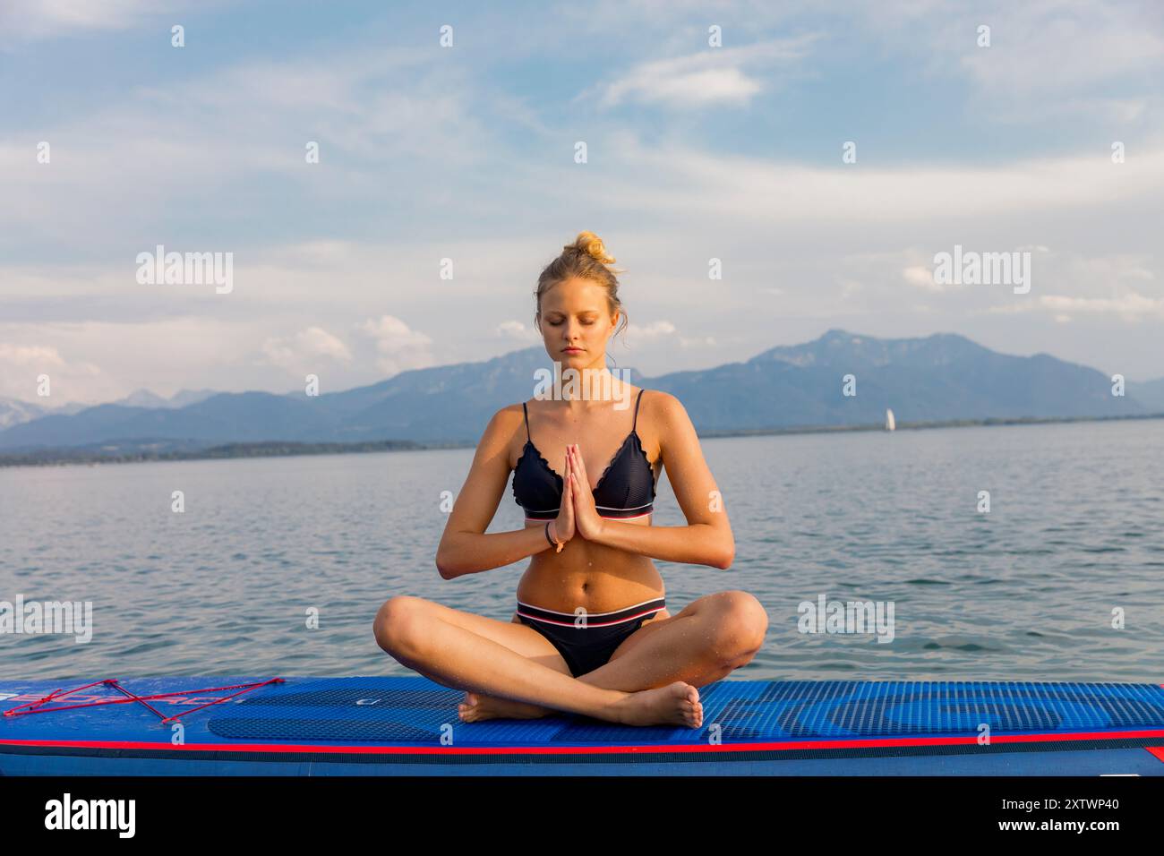Donna che medita in posizione di loto su un tappetino da yoga blu posto su una piattaforma galleggiante su un lago calmo con montagne in lontananza. Foto Stock