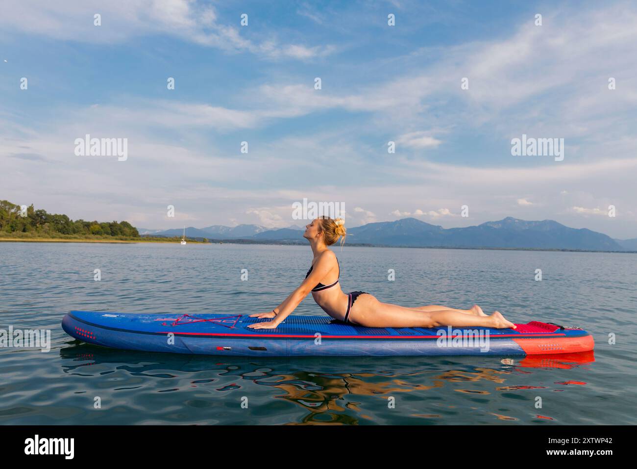 Una donna in costume da bagno gode di un momento di pace mentre è sdraiata su una tavola da paddleboard su un lago calmo con montagne sullo sfondo. Foto Stock