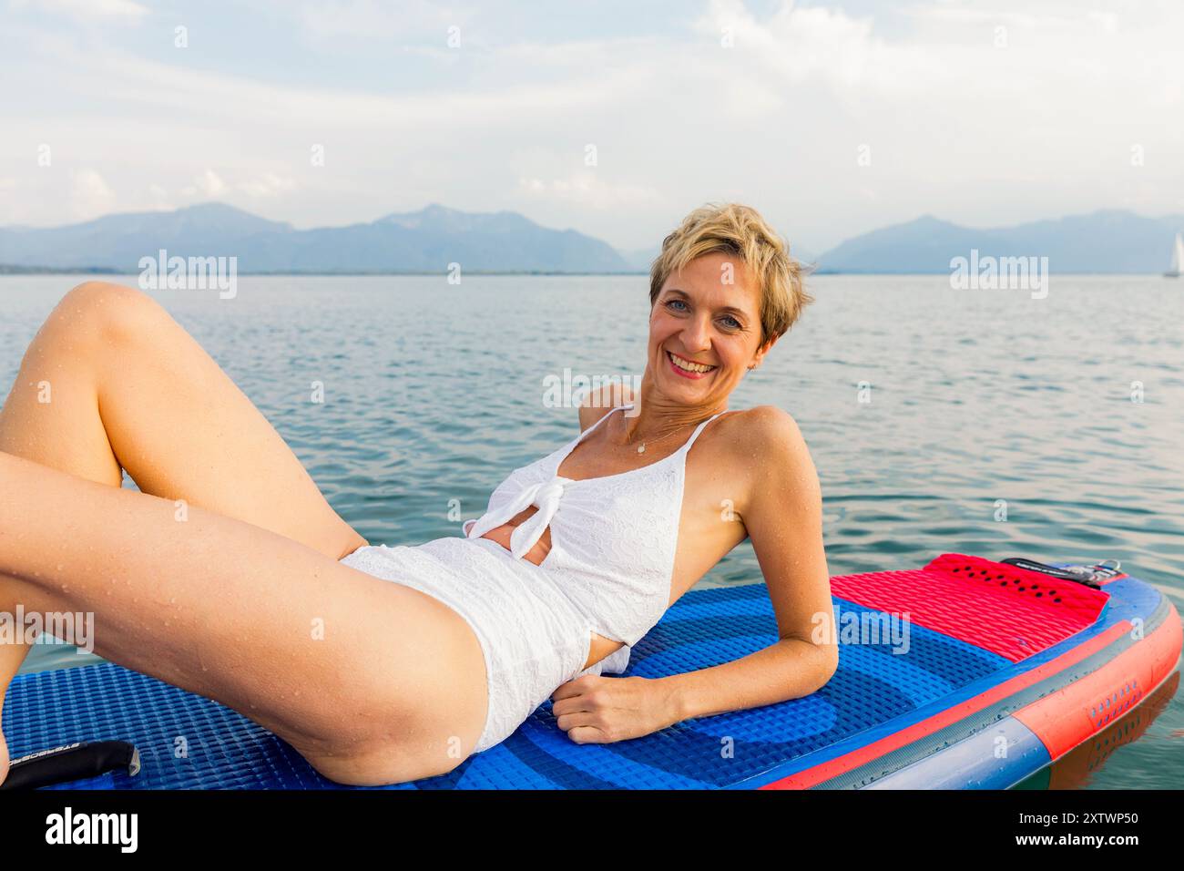 Donna sorridente che si rilassa su un paddleboard su un lago calmo con montagne in lontananza. Foto Stock