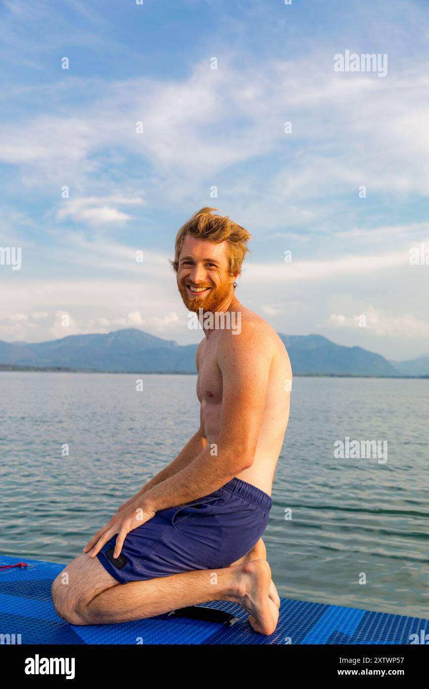 Uomo sorridente con barba inginocchiata su un tappeto blu in riva al lago con montagne sullo sfondo. Foto Stock