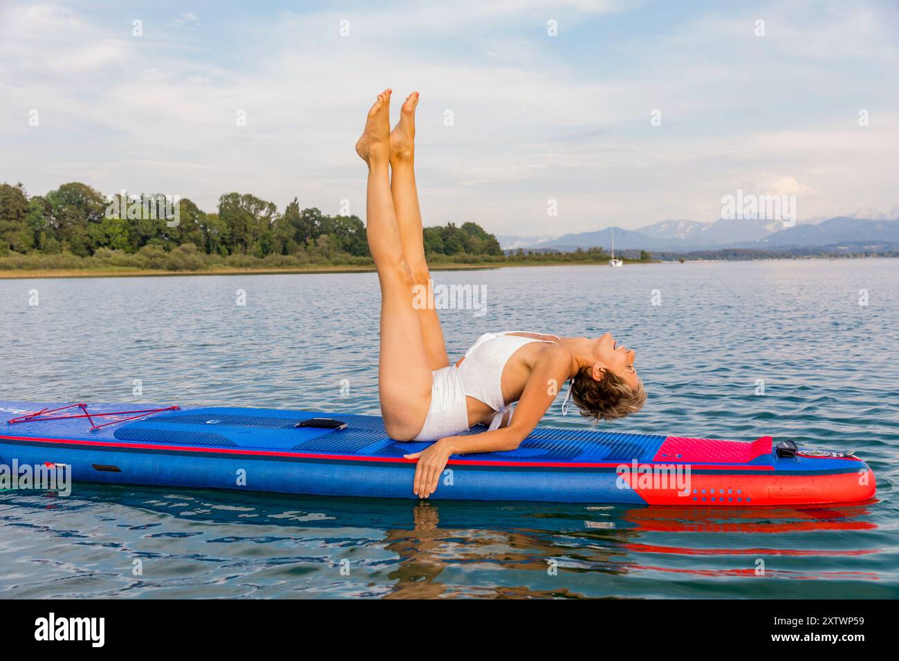 Donna che esegue yoga posa su una tavola da paddleboard su un lago calmo con montagne sullo sfondo. Foto Stock