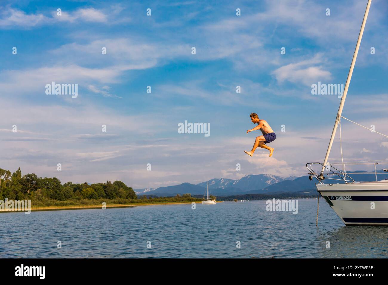 Una persona sta saltando dal lato di una barca a vela nelle calme acque blu con uno sfondo naturale panoramico e un cielo limpido. Foto Stock