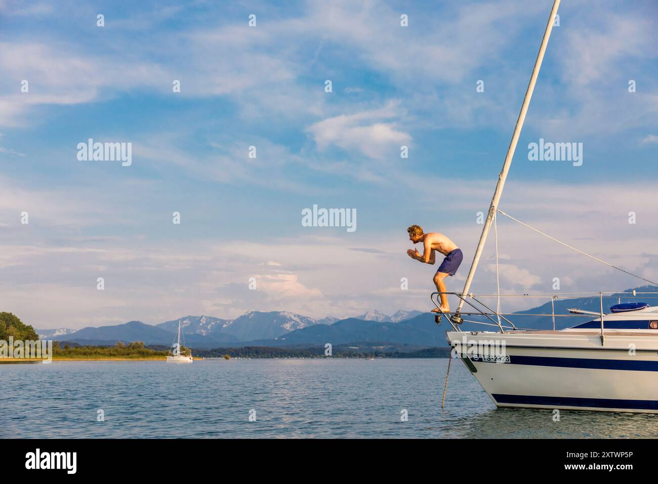 Una persona si tuffa dalla prua di una barca a vela in un tranquillo lago con montagne sullo sfondo in una giornata limpida. Foto Stock