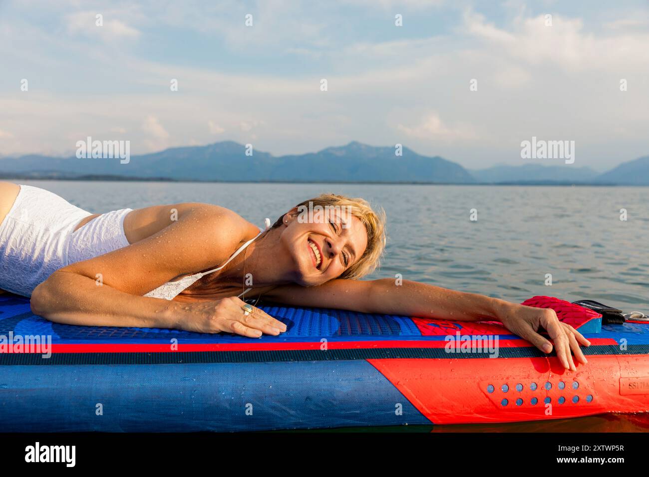 Donna sorridente sdraiata su un paddleboard che galleggia su acque calme con montagne in lontananza. Foto Stock