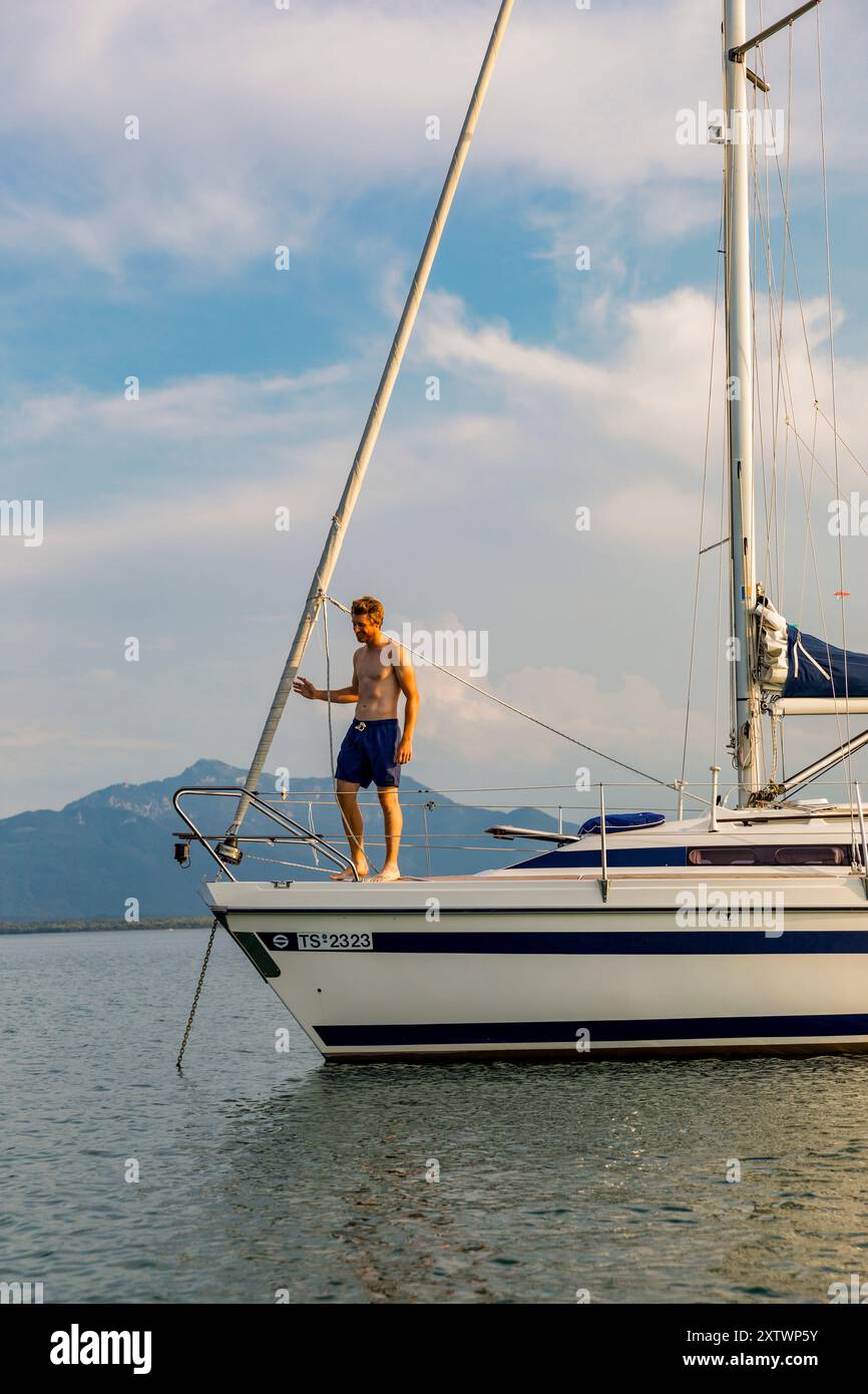 Un uomo sta sul ponte di una barca a vela bianca, tenendo le corde con un cielo limpido e un mare calmo sullo sfondo. Foto Stock