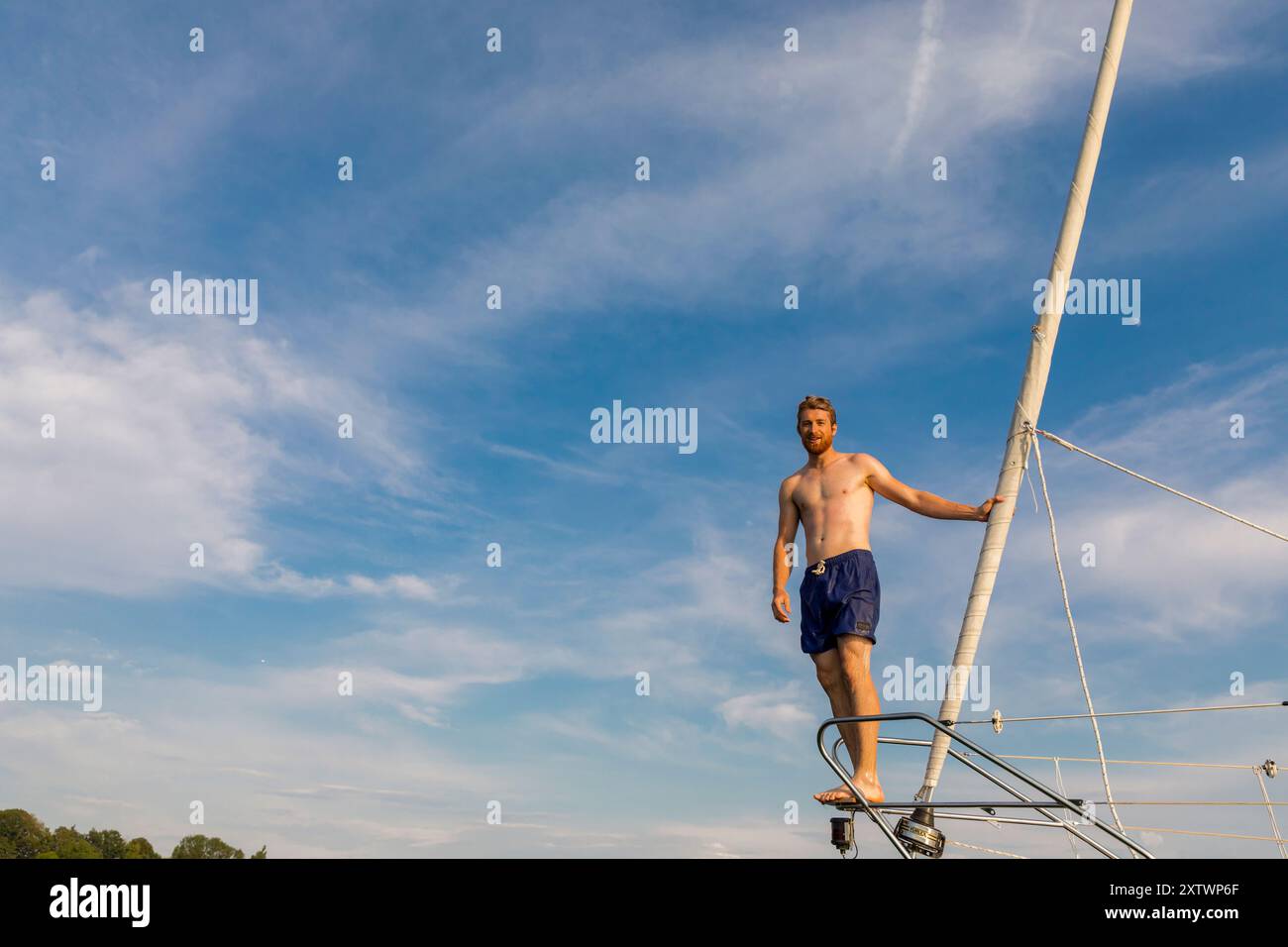 Uomo senza camicia in piedi sul ponte di una barca a vela con un cielo azzurro chiaro sullo sfondo. Foto Stock