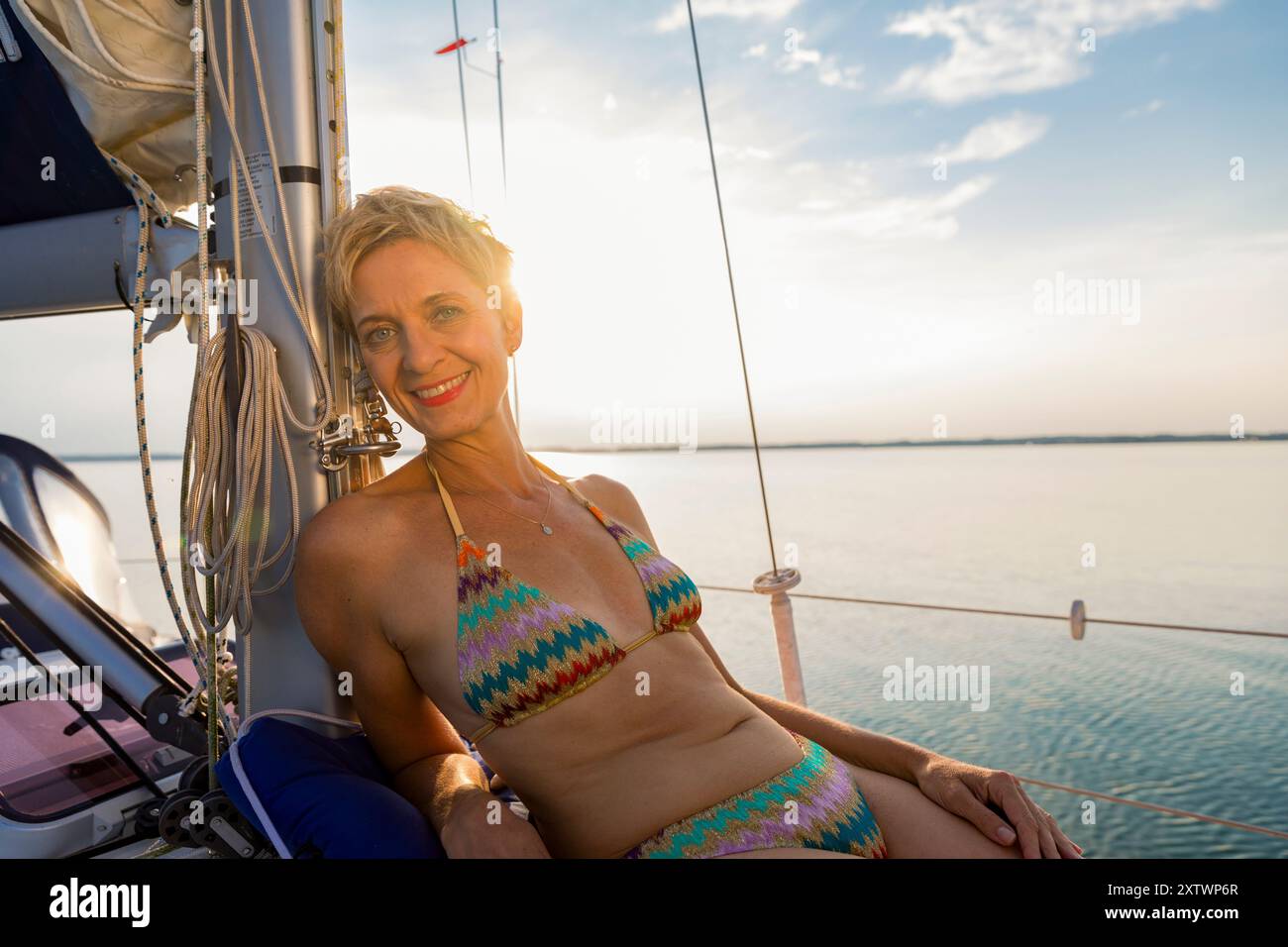 Una donna sorridente in un bikini colorato gode del sole su una barca a vela durante il tramonto. Foto Stock