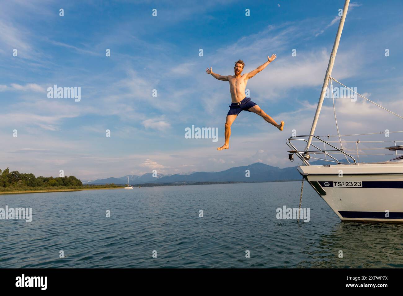 Giovane energico che salta da una barca a vela in un tranquillo lago con montagne sullo sfondo sotto un cielo azzurro. Foto Stock