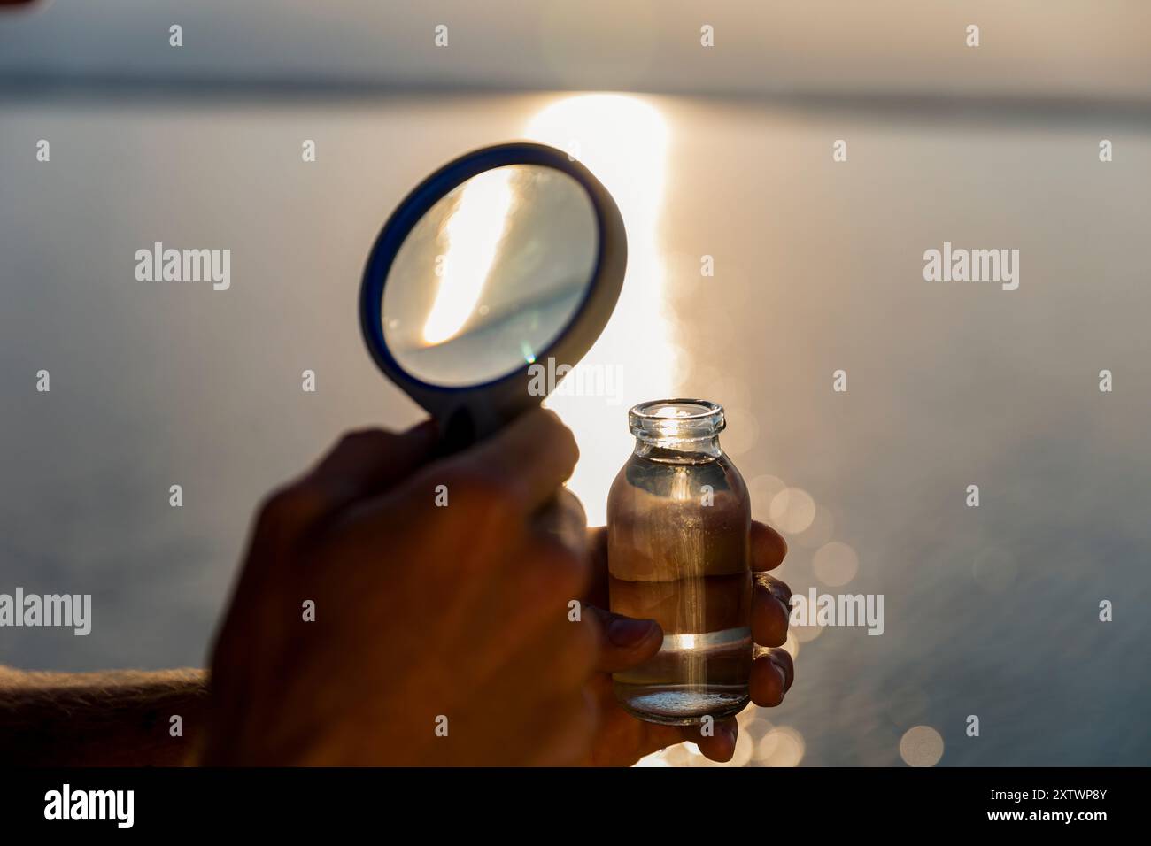 Una persona tiene in mano una lente d'ingrandimento e concentra la luce del sole su una piccola bottiglia durante un tramonto luminoso. Foto Stock