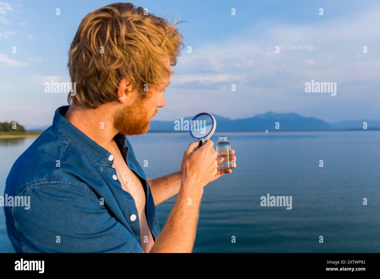 Uomo con una lente d'ingrandimento che esamina un campione d'acqua sullo sfondo di un lago panoramico Foto Stock