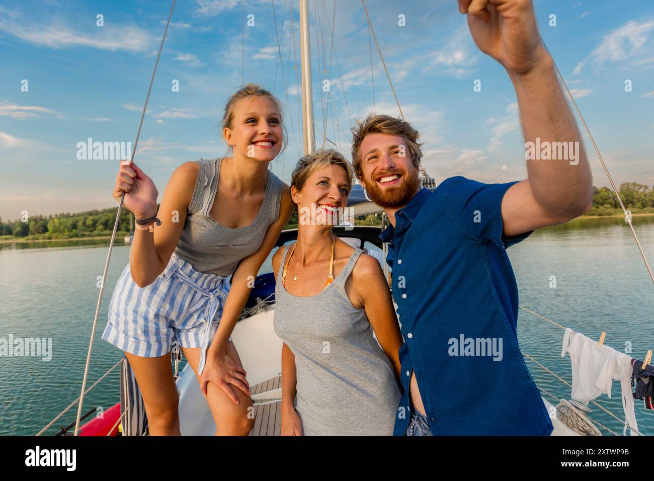 Tre adulti felici posano per una foto su una barca a vela, con un cielo azzurro limpido e un'acqua serena sullo sfondo. Foto Stock