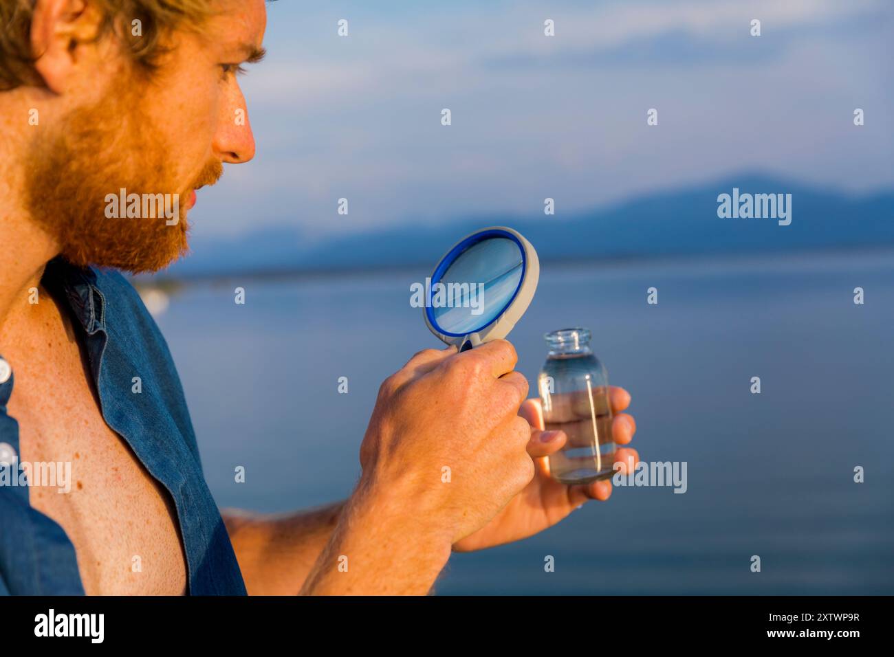 Uomo che esamina un campione d'acqua attraverso una lente di ingrandimento sullo sfondo di un lago panoramico. Foto Stock