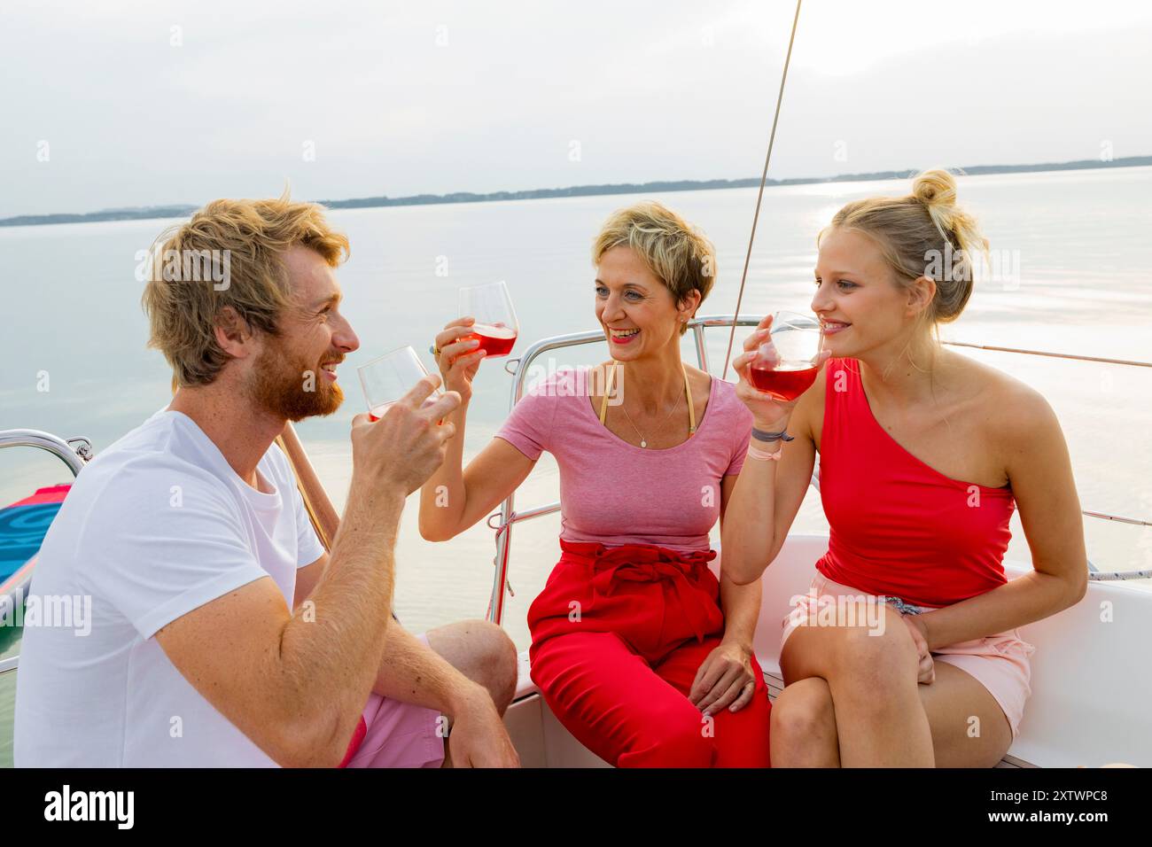 Tre amici si godono un drink e chiacchierano sul ponte di una barca a vela con un tranquillo sfondo d'acqua al tramonto. Foto Stock