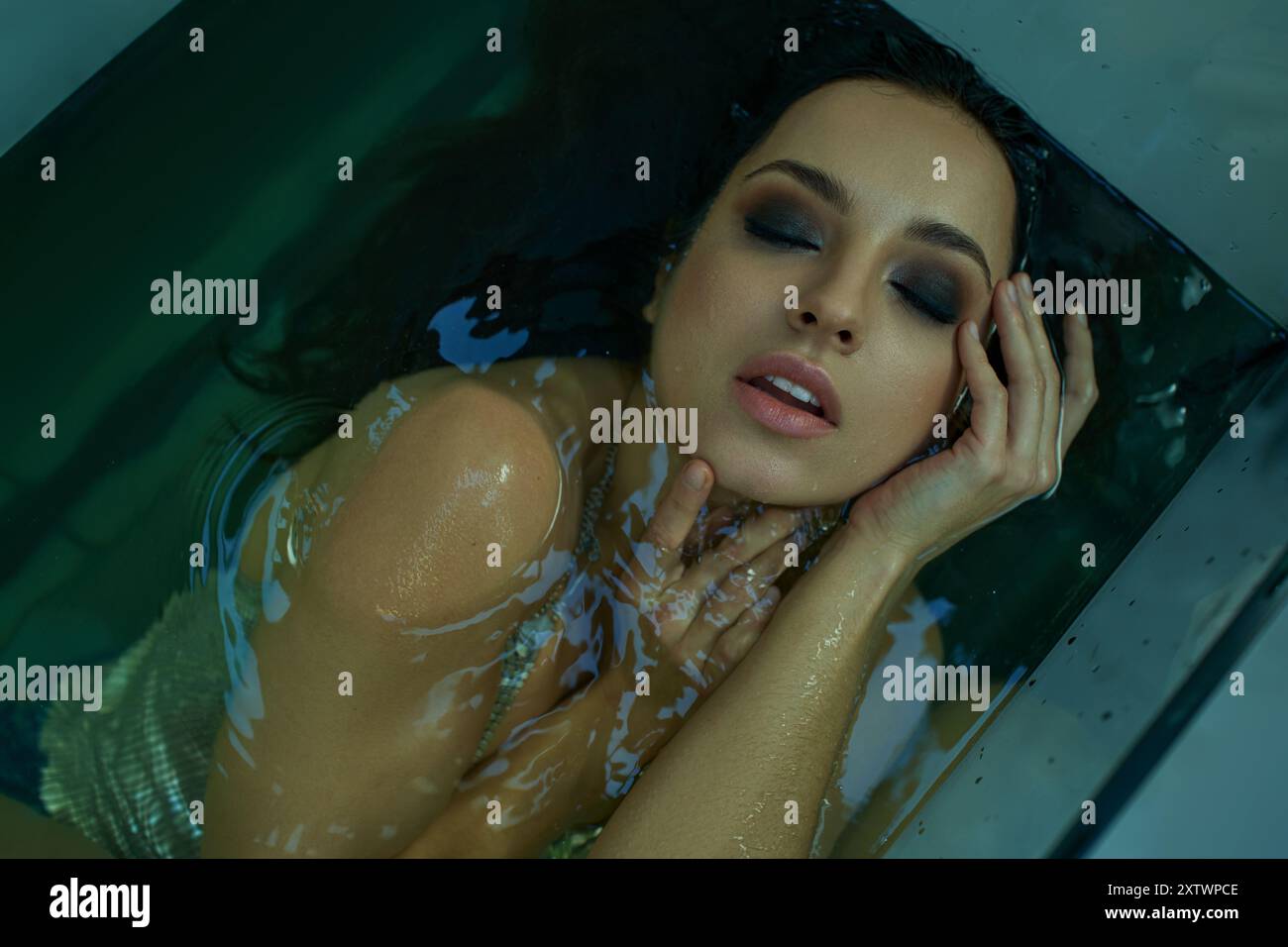 Una donna con i capelli bagnati galleggia in una vasca, gli occhi chiusi e le mani sul viso, in acqua limpida con luce soffusa per creare un'aura serena. Foto Stock