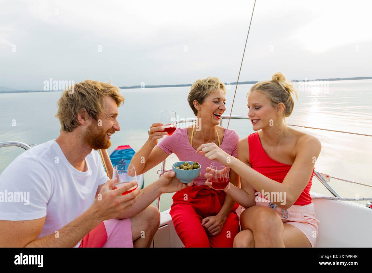 Tre amici si godono bevande e spuntini su una barca durante una giornata di sole con un cielo limpido e un lago calmo sullo sfondo. Foto Stock