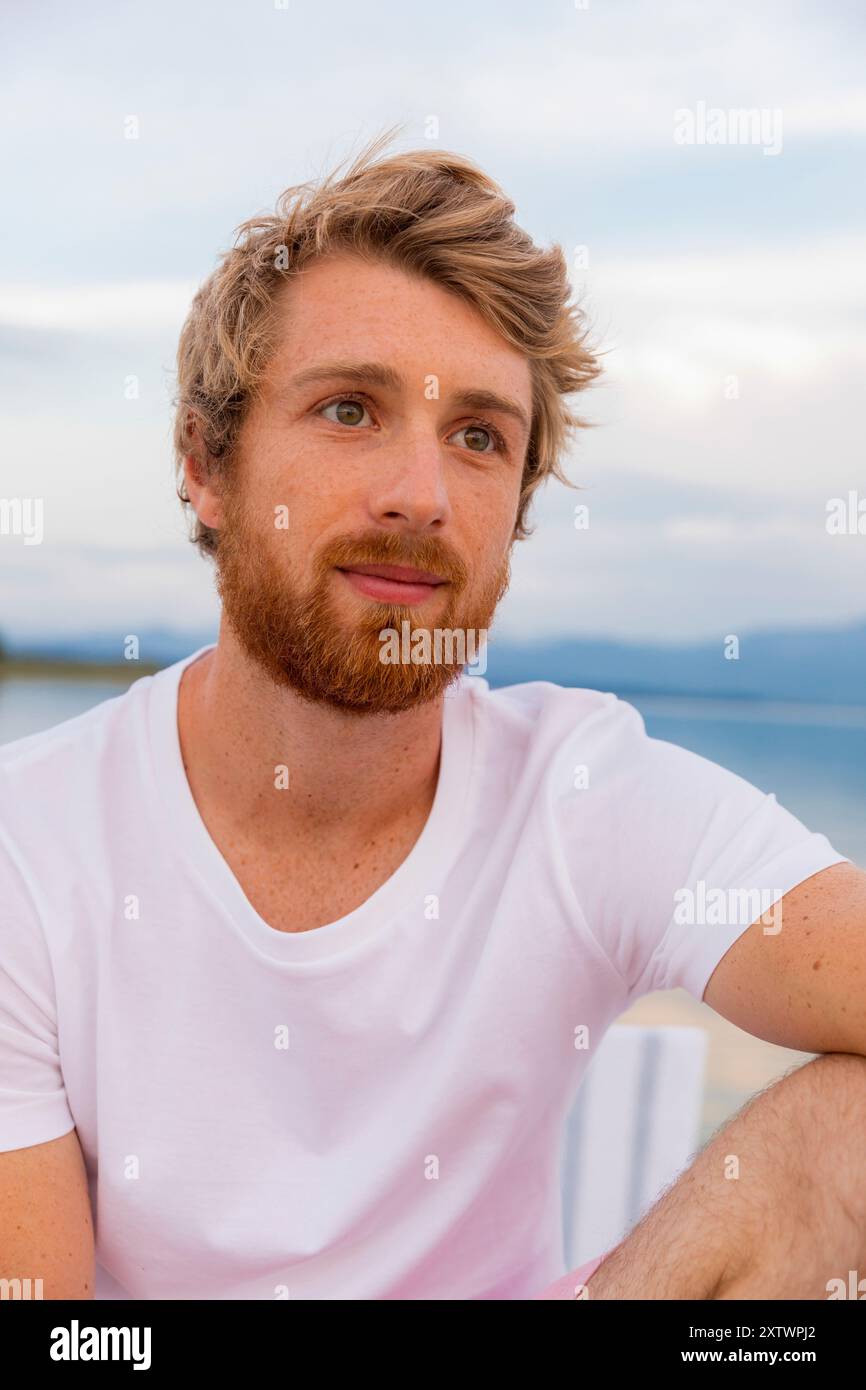 Ritratto informale di un uomo sorridente con barba e capelli in slitta con una t-shirt bianca sullo sfondo sfocato del cielo e delle montagne. Foto Stock