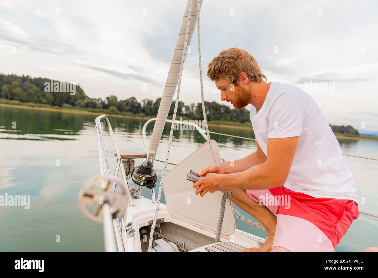 Un uomo con la barba regola l'equipaggiamento sulla poppa di una barca a vela, con acque tranquille e una costa boscosa sullo sfondo. Foto Stock