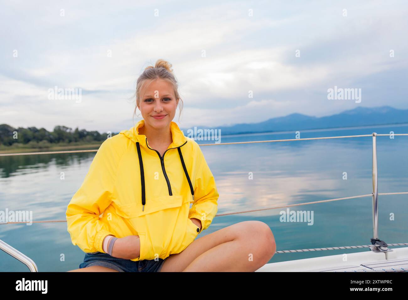 Donna in una felpa con cappuccio gialla seduta sul ponte di una barca a vela con un mare calmo e un cielo coperto sullo sfondo Foto Stock