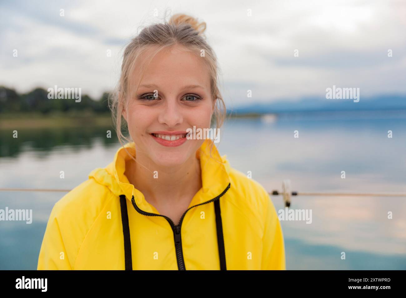 Giovane donna sorridente in una giacca gialla in piedi accanto a un lago calmo con un cielo nuvoloso sullo sfondo. Foto Stock