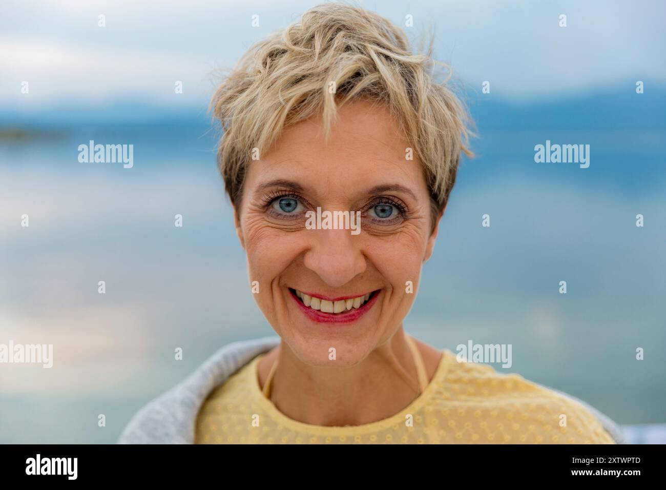 Donna sorridente con capelli biondi corti all'aperto con un lago calmo e un cielo coperto sullo sfondo. Foto Stock