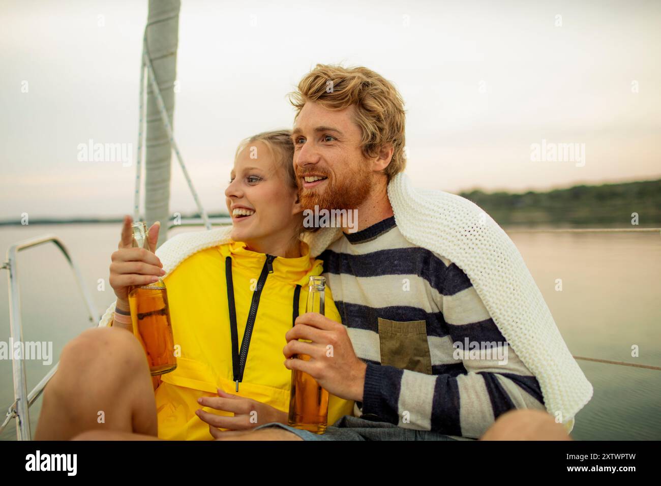 Una coppia sorridente si gode un momento insieme su una barca a vela al tramonto, tenendo in mano bottiglie di birra. Foto Stock