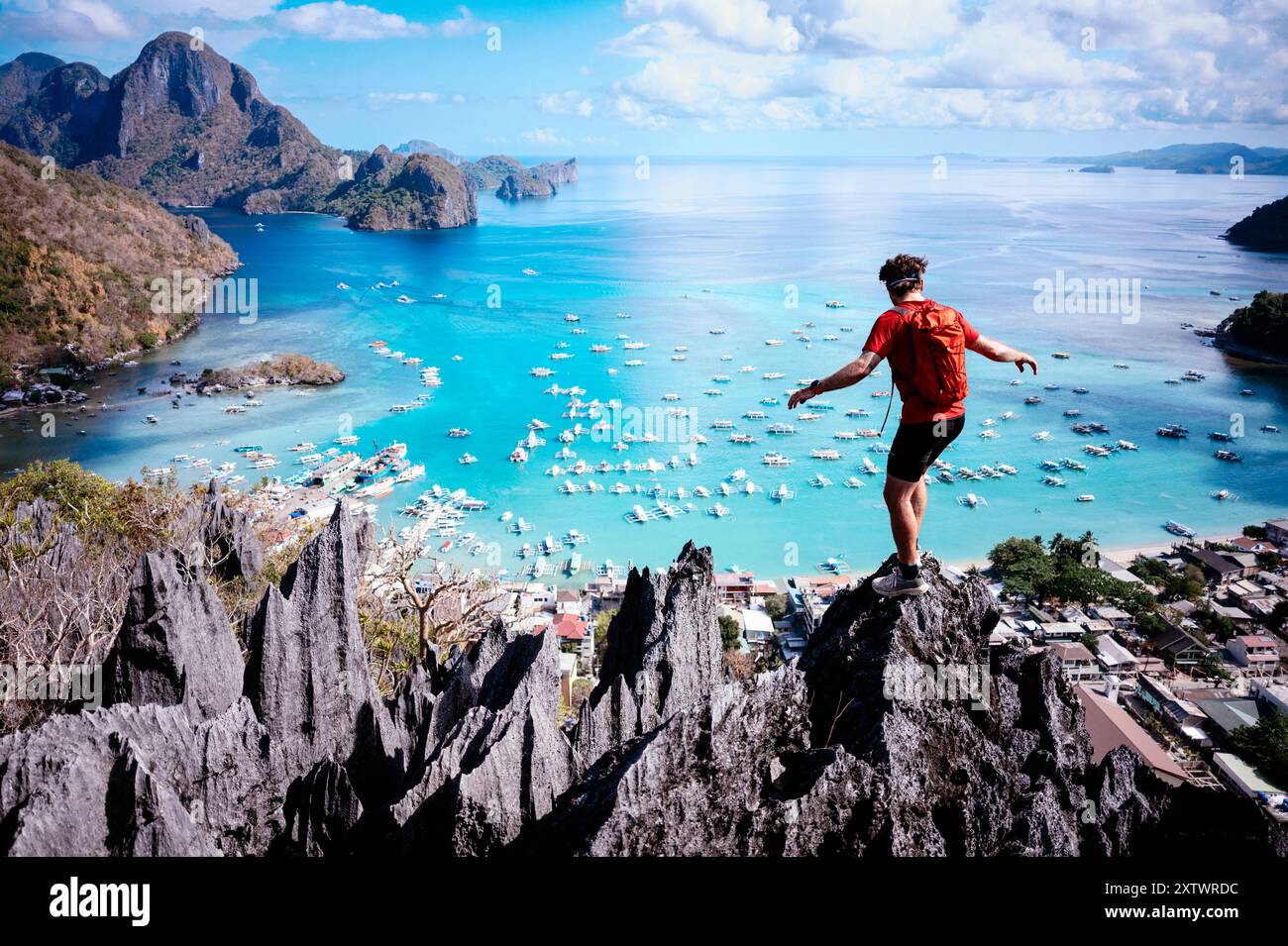 Avventuroso viaggiatore in piedi su una cima rocciosa che si affaccia su una splendida baia costiera con barche e acque cristalline. Foto Stock