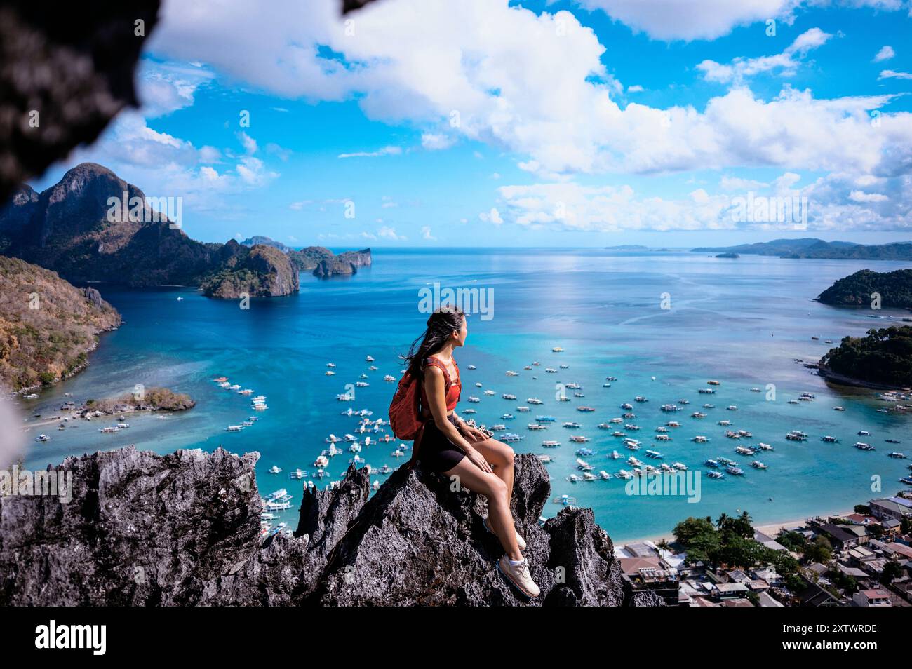 Una donna siede in cima a una cima rocciosa che si affaccia su una vista costiera mozzafiato con barche e isole sotto un cielo azzurro. Foto Stock