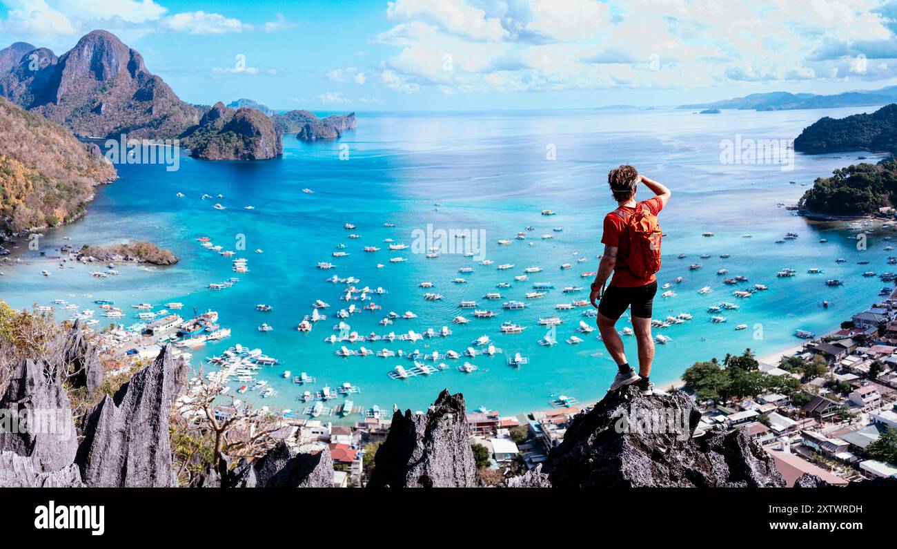 La persona si trova su una cima rocciosa che si affaccia su uno splendido mare blu disseminato di barche, delimitata da lussureggianti montagne verdi sotto un cielo limpido Foto Stock