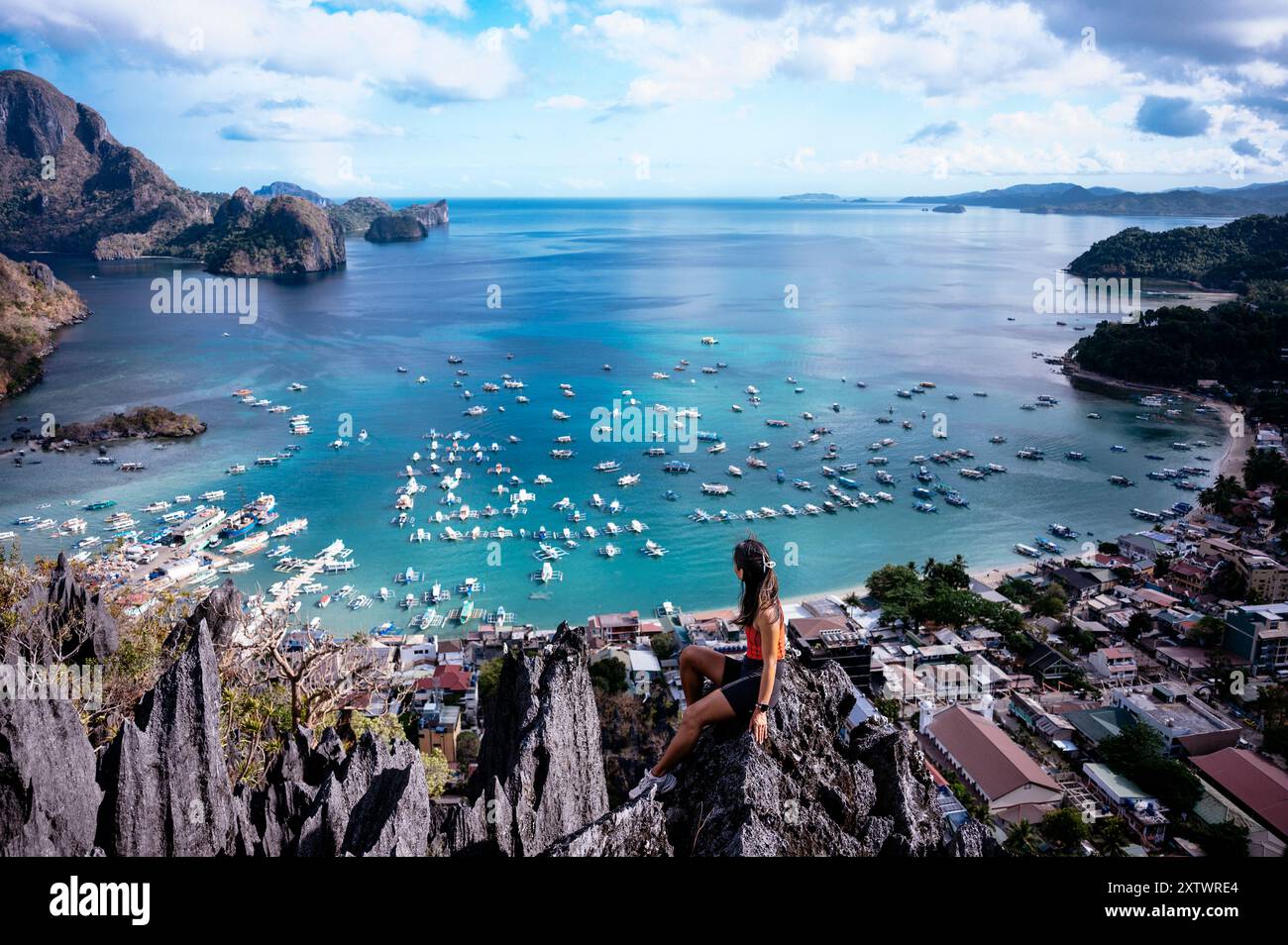 Donna seduta su un picco roccioso che si affaccia su una pittoresca baia con barche e un villaggio costiero circondato da lussureggianti colline. Foto Stock