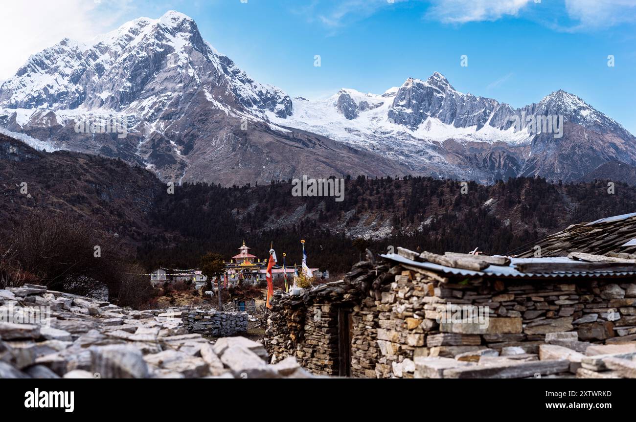 Le cime innevate sovrastano un villaggio tradizionale con case in pietra e bandiere di preghiera colorate in un tranquillo paesaggio alpino. Foto Stock