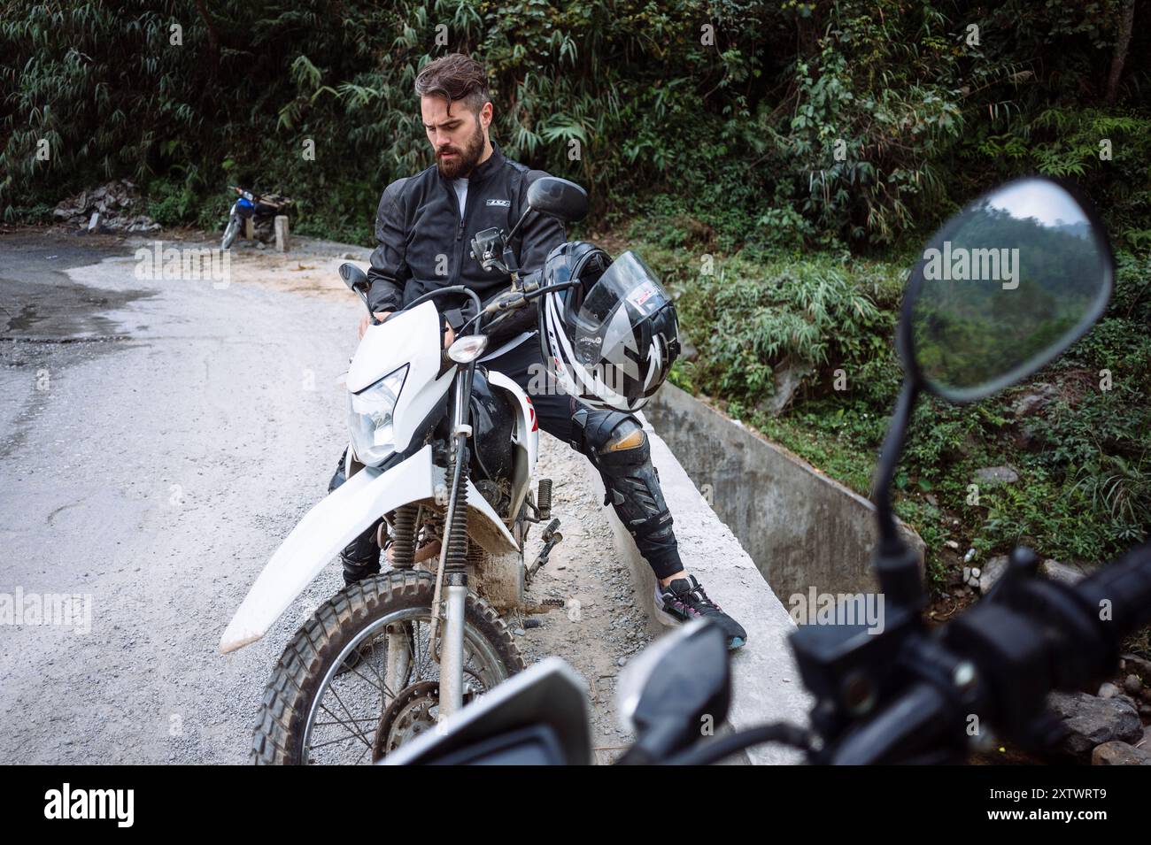 Un uomo con equipaggiamento da motocicletta riposa su una bici da terra in una strada di montagna, con uno sfondo panoramico e un altro motociclista in lontananza. Foto Stock