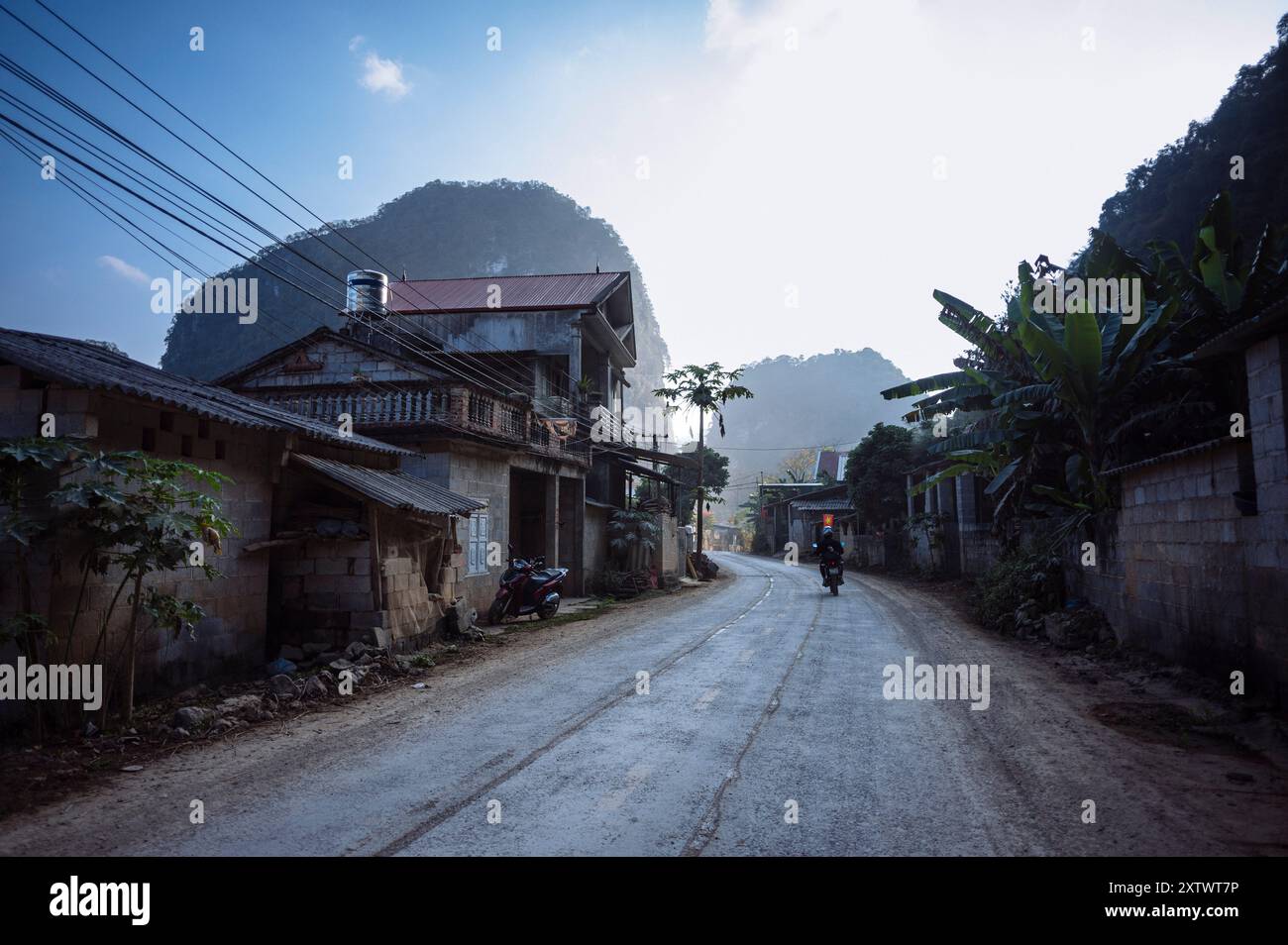 Un motociclista percorre una strada rurale polverosa fiancheggiata da edifici tradizionali e da vegetazione lussureggiante in un'area montuosa al tramonto. Foto Stock