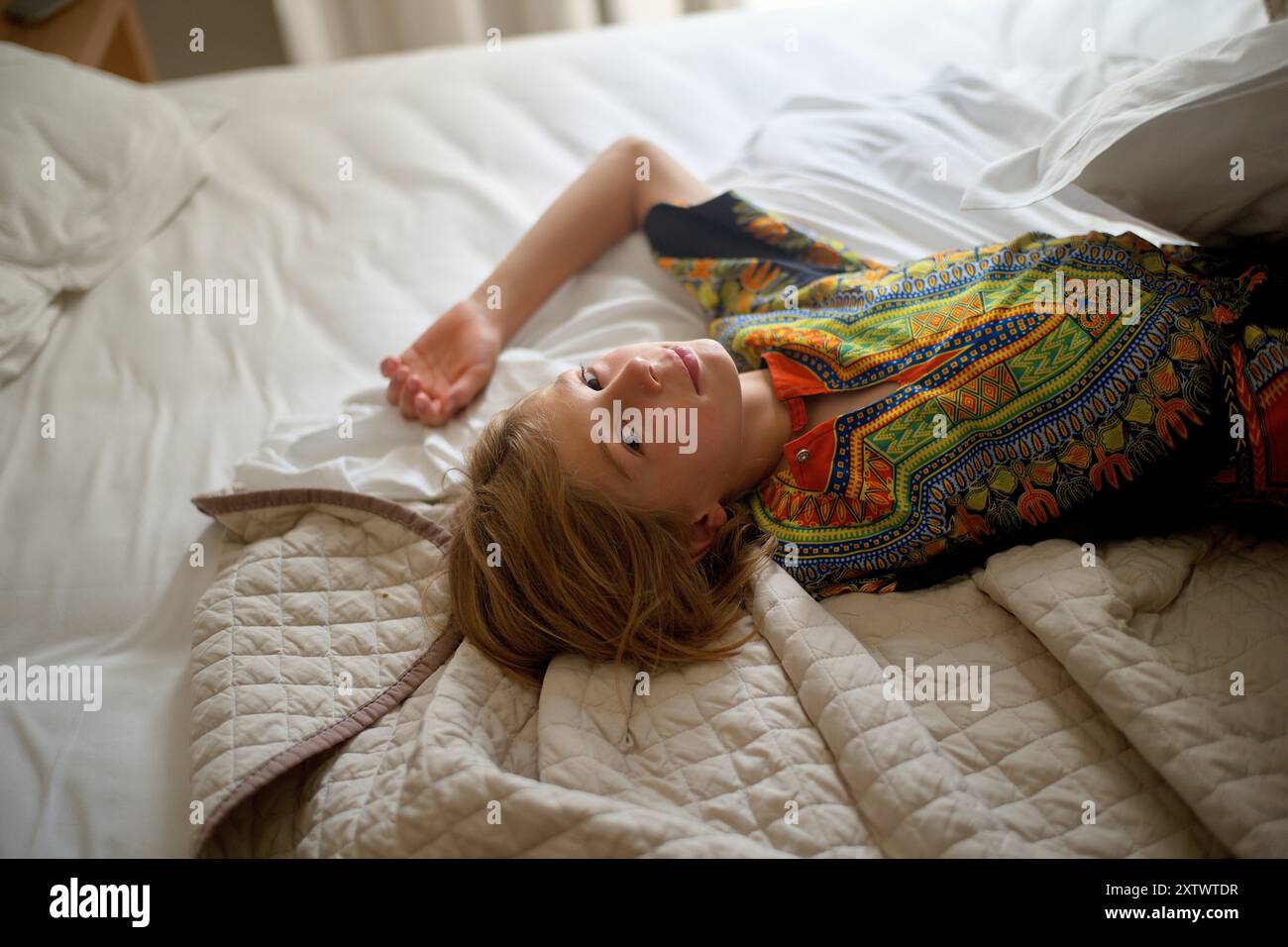 Donna sdraiata su un letto con biancheria bianca, che guarda la macchina fotografica, indossa un abito colorato. Foto Stock Donna sdraiata su un letto con biancheria bianca, che guarda la macchina fotografica, indossa un abito colorato. Foto Stock