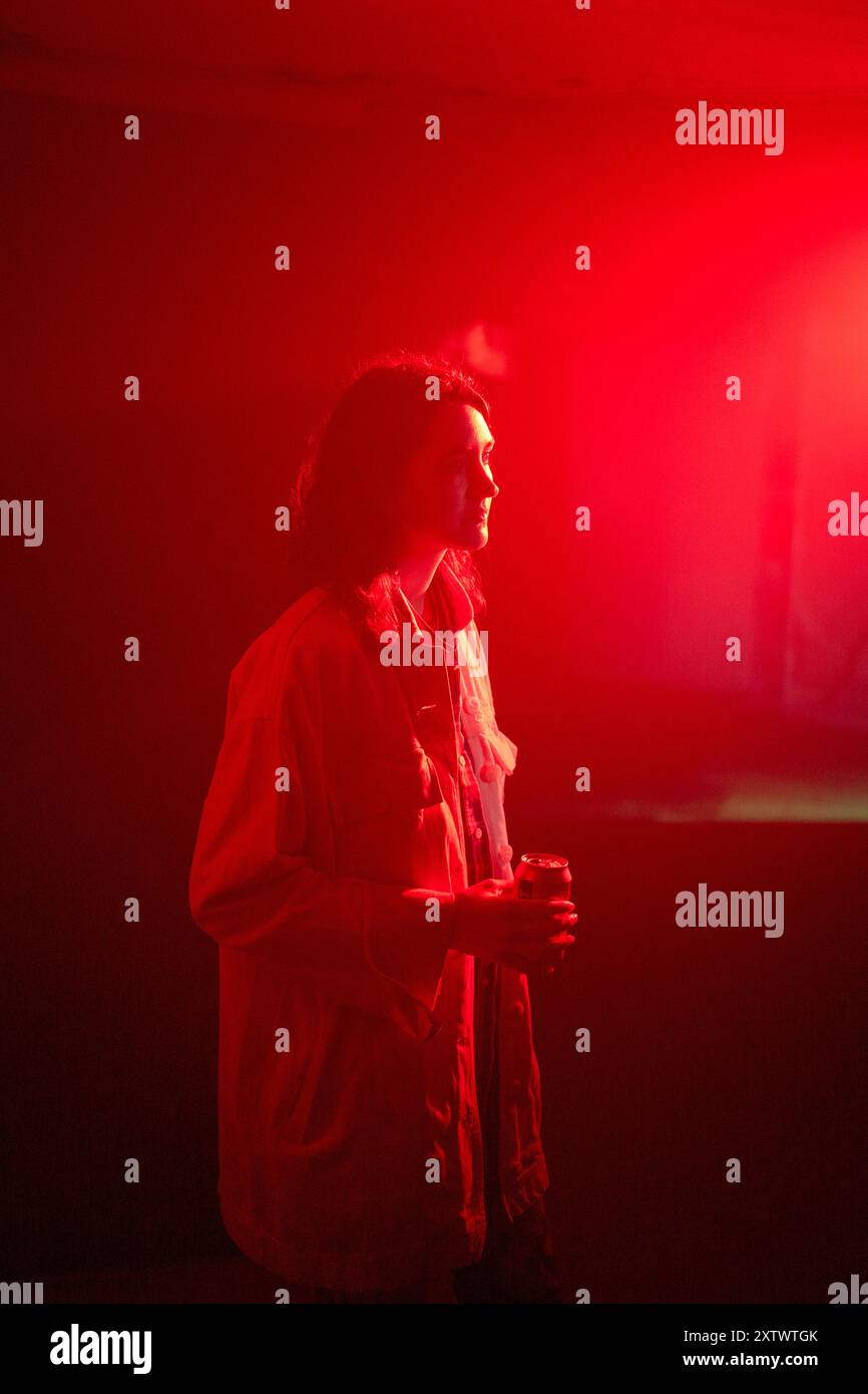 Una donna si trova in una stanza illuminata di rosso che tiene un drink, guardando in prospettiva di lato. Foto Stock