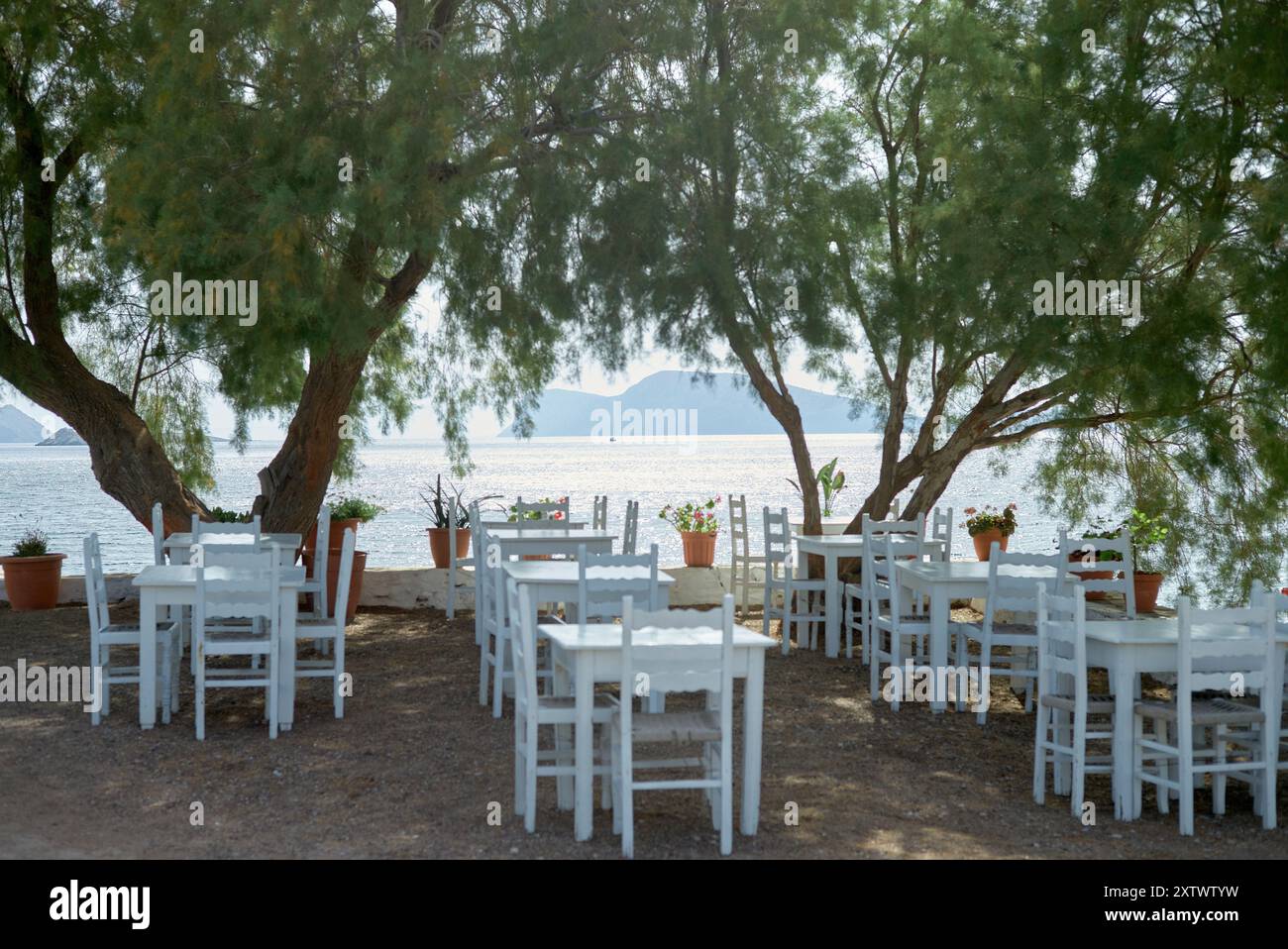 Ristorante sul mare con sedie bianche e tavoli all'ombra degli alberi, affacciato su una baia tranquilla. Foto Stock