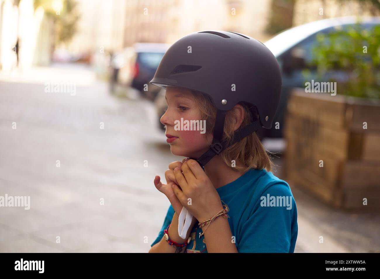 Un bambino con i capelli lunghi che indossa un casco fissa il cinturino sotto il mento in una strada cittadina. Foto Stock