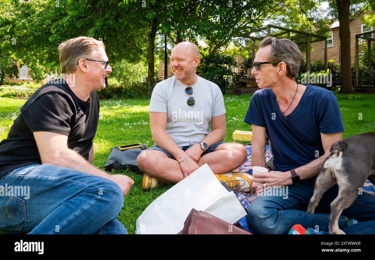 Tre uomini seduti su una coperta da picnic in un parco che si godono una conversazione con un cane nelle vicinanze. Foto Stock