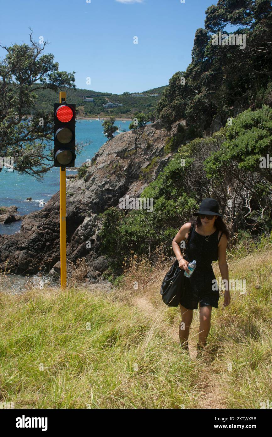 Una donna con un abito nero e un cappello cammina vicino a un semaforo su una collina erbosa vicino al mare, Waiheke Island, nuova Zelanda Foto Stock