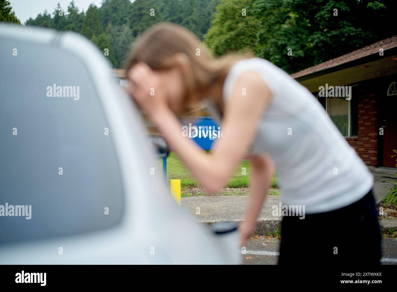 Una donna che si appoggia per guardare il motore della sua auto con un'espressione frustrata fuori da un edificio con il segno della Divisione." Foto Stock