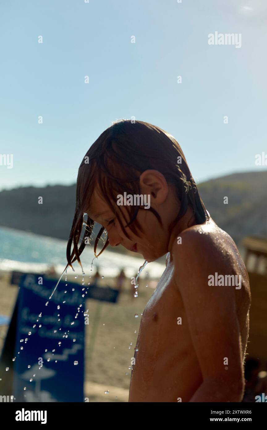 Un ragazzino immerso in gocce d'acqua si erge su una spiaggia soleggiata con il mare e le sdraio sullo sfondo. Foto Stock